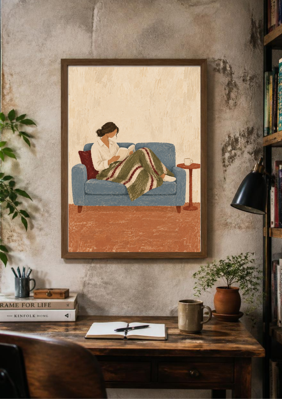 A framed art print depicting a woman reading on a blue couch, hung on a textured concrete wall above a wooden desk with books, a mug, and a plant.